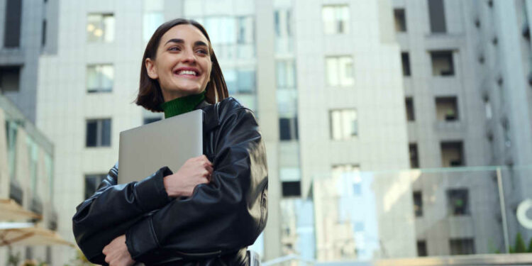 smiling woman holding laptop in front of buildings.